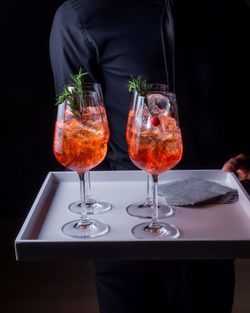 Four glasses of orange cocktails garnished with rosemary, served on a white tray held by a waiter in a dark outfit.
