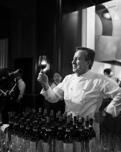 Chef in white uniform examining a glass of red wine behind a bar with numerous wine bottles at a catering event.