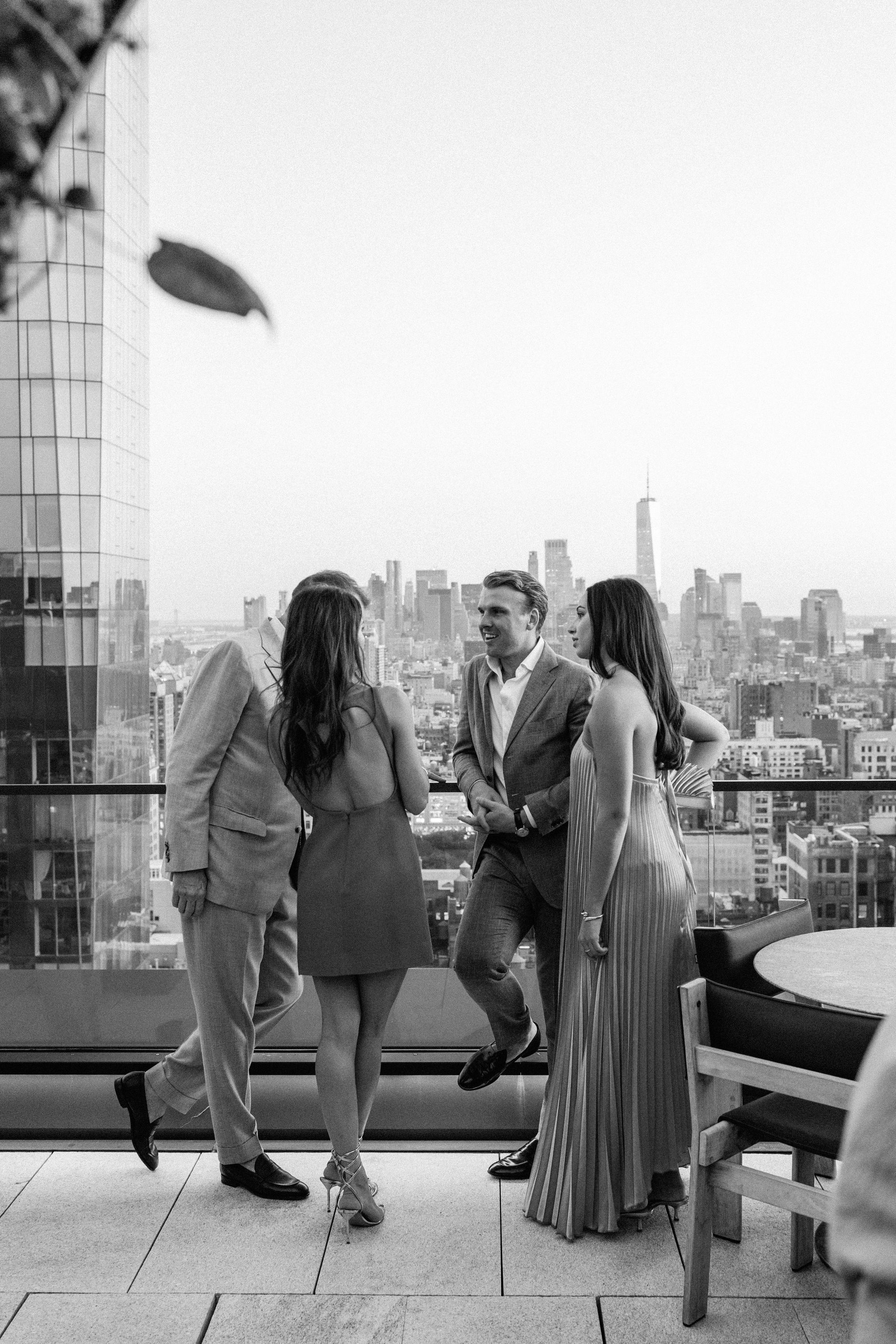 Guests enjoy a gathering with a panoramic view of New York City, showcasing elegant attire and a lively atmosphere.