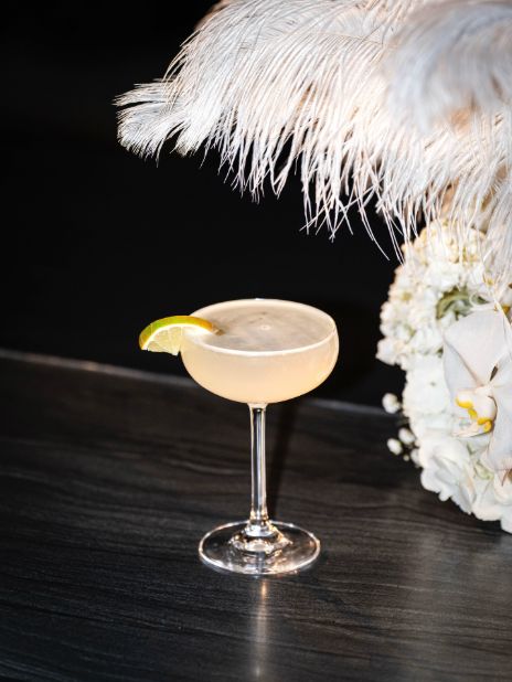 A cocktail garnished with a lime slice in a coupe glass, placed on a black surface with decorative feathers.