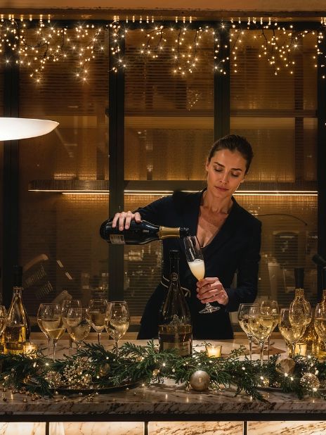 A bartender pours champagne into a glass at a decorated event bar with festive lights and greenery.