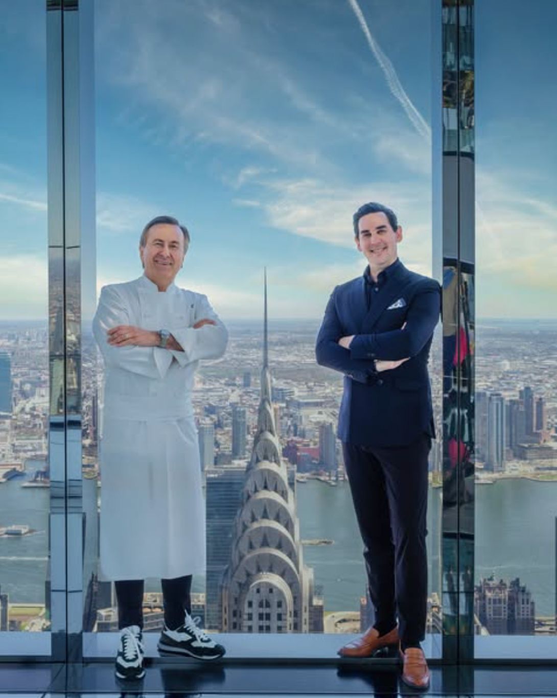 Two chefs pose with the New York City skyline in the background, featuring iconic buildings like the Chrysler Building.